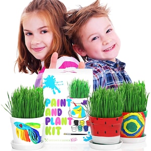 May include: Two children hold up a box with the text "Paint and Plant Kit" and a picture of a unicorn pot with green grass growing in it. There are three pots with green grass growing in them, one with a rainbow fish design, one with a watermelon design, and one with a rainbow design. The pots are white with colorful designs.
