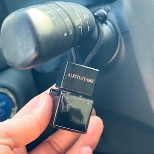 Può includere: Un deodorante per auto nero e quadrato con la scritta "ALITTLE TAME" sulla parte superiore. Il deodorante è appeso a un cordino nero attaccato alla leva degli indicatori di direzione di un'auto. L'interno dell'auto è visibile sullo sfondo.