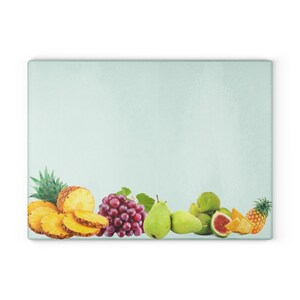 Puede incluir: Una tabla de cortar de vidrio azul claro con un borde de fruta. La fruta incluye rodajas de piña, uvas, peras, higos y trozos de piña.
