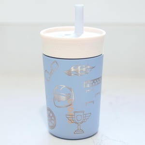 May include: A light blue tumbler with a cream-colored lid and a matching straw. The exterior features a repeating pattern of silver-toned racing-themed icons, including a helmet, a trophy, a checkered flag, a wheel, and flame designs.