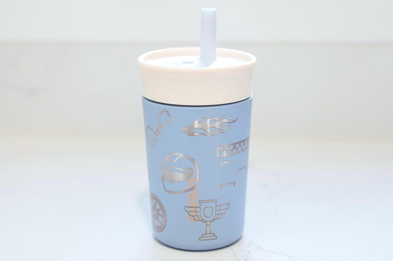 May include: A light blue tumbler with a cream-colored lid and a matching straw. The exterior features a repeating pattern of silver-toned racing-themed icons, including a helmet, a trophy, a checkered flag, a wheel, and flame designs.