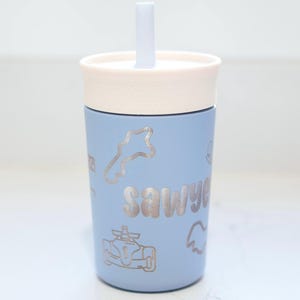 May include: A light blue tumbler with a cream-colored lid and a matching straw. The cup features silver-toned line art graphics of a race car, a track outline, and the name Sawyer printed in a bold, rounded font on the front surface.