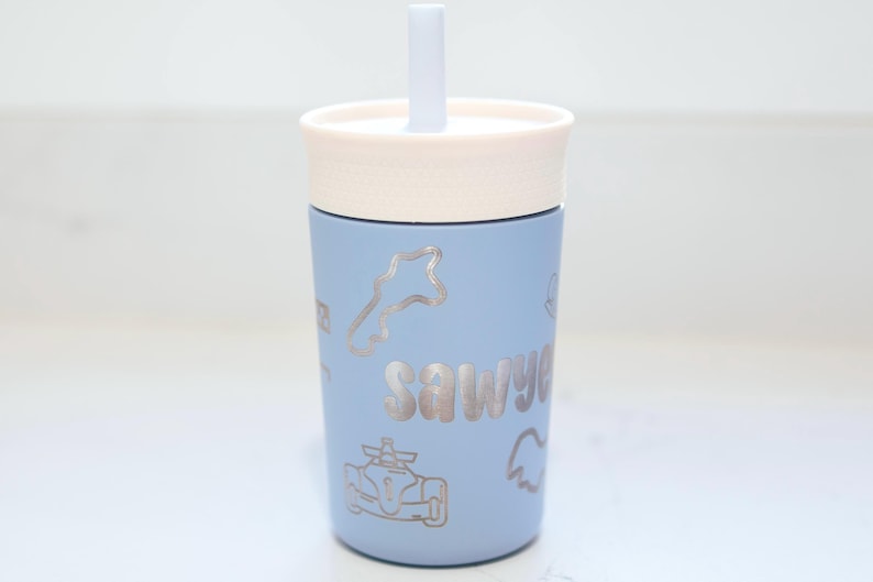 May include: A light blue tumbler with a cream-colored lid and a matching straw. The cup features silver-toned line art graphics of a race car, a track outline, and the name Sawyer printed in a bold, rounded font on the front surface.