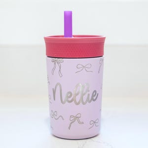 May include: A light purple tumbler with a pink lid and a purple straw. The tumbler features a repeating pattern of thin silver bows and the name Nellie written in a silver cursive script across the center of the cup's exterior surface.