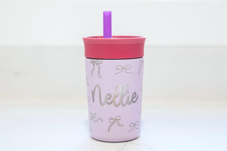 May include: A light purple tumbler with a pink lid and a purple straw. The tumbler features a repeating pattern of thin silver bows and the name Nellie written in a silver cursive script across the center of the cup's exterior surface.