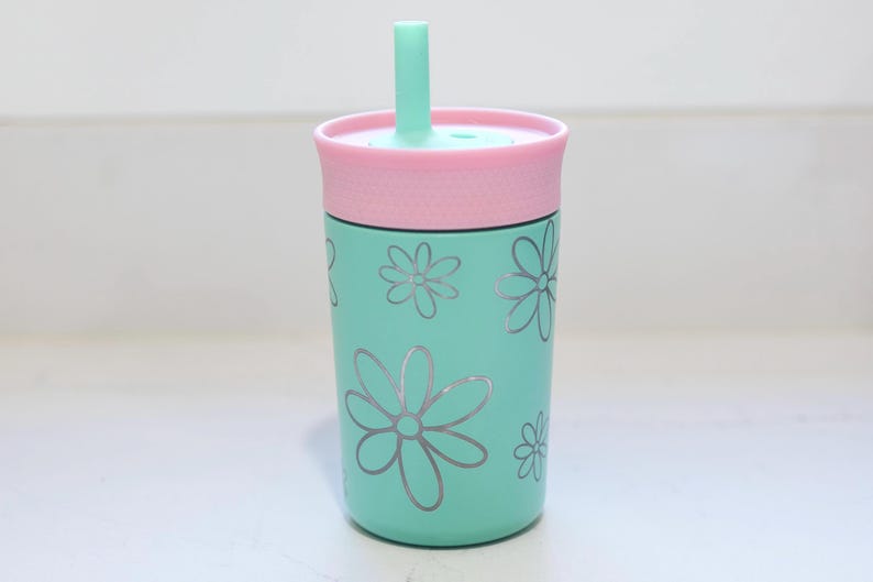 May include: A mint green tumbler featuring a repeating pattern of silver-colored flower outlines. The cup includes a light pink lid with a textured band and a matching mint green straw protruding from the center of the lid top.