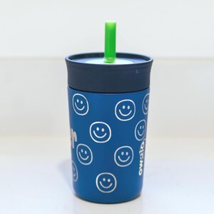 May include: A blue tumbler with a dark blue lid and a bright green straw. The exterior of the cup features a repeating pattern of white line-drawn smiley faces. The word Owala is printed vertically in white on the side of the blue cup body.