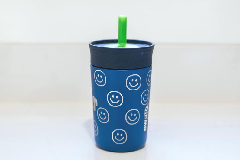 May include: A blue tumbler with a dark blue lid and a bright green straw. The exterior of the cup features a repeating pattern of white line-drawn smiley faces. The word Owala is printed vertically in white on the side of the blue cup body.