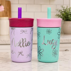 May include: Two insulated tumblers with lids and straws. The left tumbler is light purple with silver bow graphics and the name Nellie. The right tumbler is mint green with silver flower graphics and the name Lucy. Both have textured lids.