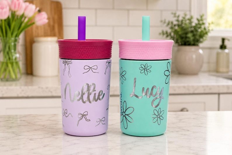 May include: Two insulated tumblers with lids and straws. The left tumbler is light purple with silver bow graphics and the name Nellie. The right tumbler is mint green with silver flower graphics and the name Lucy. Both have textured lids.