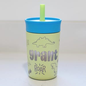 May include: A light green tumbler with a blue lid and a light green straw. The tumbler features the name grant in dark grey letters, surrounded by dark grey line art illustrations of dinosaurs and the word ROAR in a stylized font.