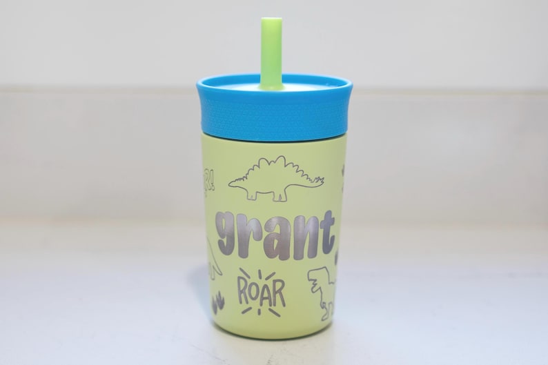 May include: A light green tumbler with a blue lid and a light green straw. The tumbler features the name grant in dark grey letters, surrounded by dark grey line art illustrations of dinosaurs and the word ROAR in a stylized font.