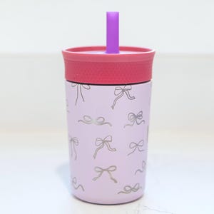 May include: A light purple tumbler featuring a repeating pattern of thin, metallic silver bows. The cup has a textured dark pink lid and a solid purple straw protruding from the center. The design is consistent across the cylindrical body.
