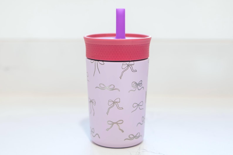 May include: A light purple tumbler featuring a repeating pattern of thin, metallic silver bows. The cup has a textured dark pink lid and a solid purple straw protruding from the center. The design is consistent across the cylindrical body.