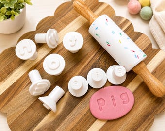 May include: A white rolling pin with colourful sprinkles, several white circular letter stamps, and a pink oval piece of dough with the word pig embossed on it. The stamps feature individual letters, and the rolling pin has wooden handles.