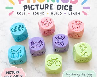 May include: A set of seven colourful dice featuring embossed phonics-related imagery including a sled, map, dog, drum, pond, person jumping, bicycle, and kite. The text PHONICS PICTURE DICE ROLL SOUND BUILD LEARN is displayed at the top of the image.