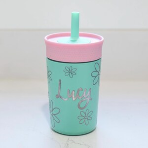 May include: A mint green tumbler with a pink lid and a matching mint green straw. The tumbler features a silver-colored floral pattern and the name Lucy written in a cursive script font across the center of the cup's exterior surface.