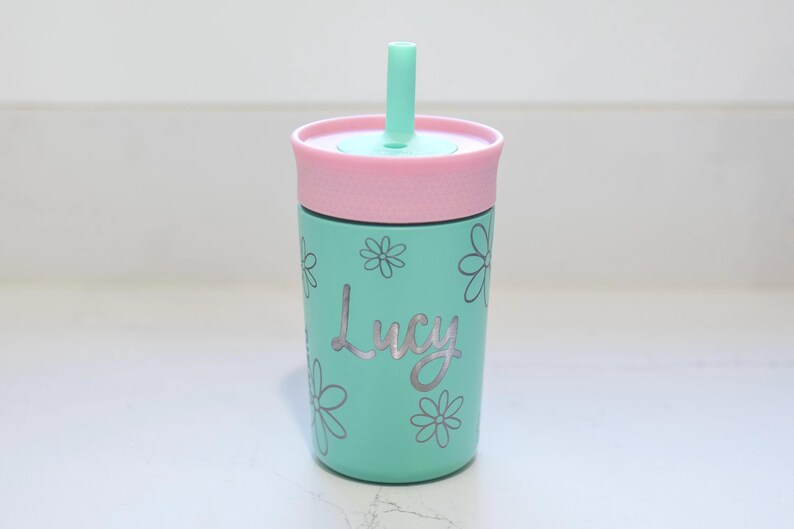 May include: A mint green tumbler with a pink lid and a matching mint green straw. The tumbler features a silver-colored floral pattern and the name Lucy written in a cursive script font across the center of the cup's exterior surface.