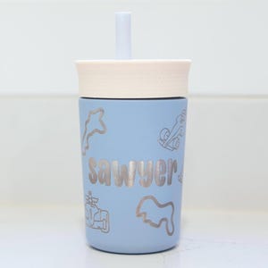 May include: A light blue tumbler with a cream-colored lid and a matching straw. The cup features the name "sawyer" in a bold, centered font, surrounded by various engraved-style line art illustrations of race cars and track outlines.