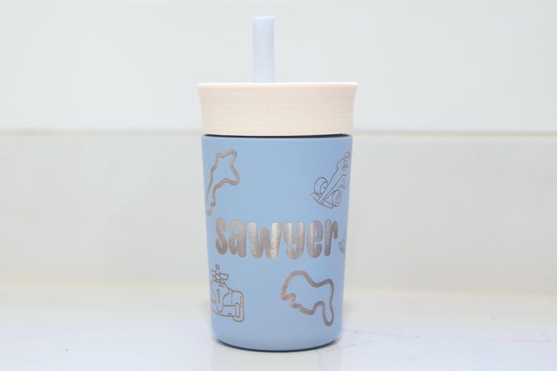 May include: A light blue tumbler with a cream-colored lid and a matching straw. The cup features the name "sawyer" in a bold, centered font, surrounded by various engraved-style line art illustrations of race cars and track outlines.