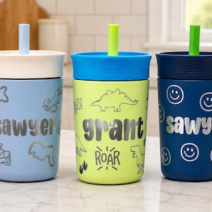 May include: Three plastic tumblers with lids and straws. The light blue cup features the name "sawyer" and race car graphics. The lime green cup features the name "grant," dinosaur graphics, and the word "ROAR." The navy cup features the name "sawyer" and smiley face graphics.
