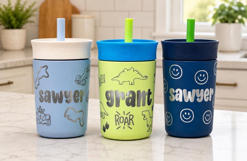 May include: Three plastic tumblers with lids and straws. The light blue cup features the name "sawyer" and race car graphics. The lime green cup features the name "grant," dinosaur graphics, and the word "ROAR." The navy cup features the name "sawyer" and smiley face graphics.