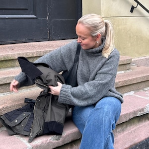 May include: A woman wearing a gray turtleneck sweater and blue jeans sits on stone steps. She is holding a dark green and black jacket. She is also wearing brown loafers.