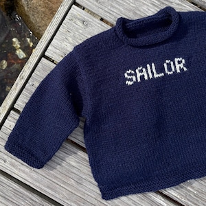 May include: A navy blue long-sleeved knit jumper featuring a rolled collar and the word SAILOR knitted in white yarn across the chest. The garment has a textured stitch pattern throughout the body and sleeves, with a simple hem at the waist.