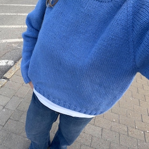 May include: A bright blue long-sleeved knit jumper with a ribbed crew neckline and hem, worn over a white undershirt, paired with blue denim jeans and dark brown leather shoes. The jumper features a chunky knit texture and raglan sleeves.