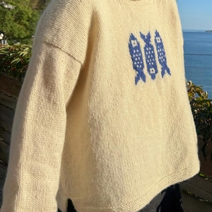 K&ouml;nnte beinhalten: Ein cremefarbener, lang&auml;rmeliger Strickpullover mit Rundhalsausschnitt und gerolltem Kragenrand. Auf der Vorderseite des Pullovers befindet sich ein zentriertes Grafikmuster aus drei vertikal ausgerichteten Fischen, die in Dunkelblau eingestrickt sind.