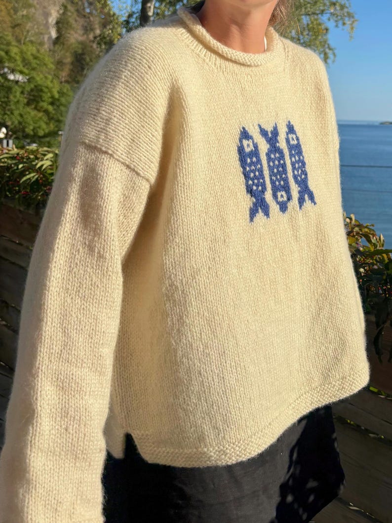 K&ouml;nnte beinhalten: Ein cremefarbener, lang&auml;rmeliger Strickpullover mit Rundhalsausschnitt und gerolltem Kragenrand. Auf der Vorderseite des Pullovers befindet sich ein zentriertes Grafikmuster aus drei vertikal ausgerichteten Fischen, die in Dunkelblau eingestrickt sind.