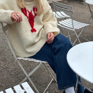 K&ouml;nnte beinhalten: Ein cremefarbener, lang&auml;rmeliger Strickpullover mit einer auff&auml;lligen roten Hummergrafik auf der Brust, kombiniert mit einer dunkelblauen Jeans, wei&szlig;en Socken und schwarzen Slippern. Der Pullover hat einen gerippten Halsausschnitt.