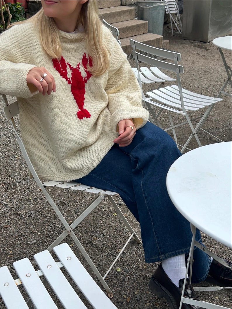 K&ouml;nnte beinhalten: Ein cremefarbener, lang&auml;rmeliger Strickpullover mit einer auff&auml;lligen roten Hummergrafik auf der Brust, kombiniert mit einer dunkelblauen Jeans, wei&szlig;en Socken und schwarzen Slippern. Der Pullover hat einen gerippten Halsausschnitt.