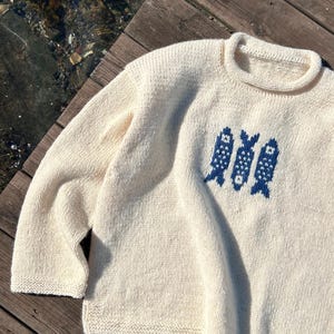 K&ouml;nnte beinhalten: Ein cremefarbener, lang&auml;rmeliger Strickpullover mit Rundhalsausschnitt und einem dunkelblauen Grafikmuster aus drei stilisierten Fischen auf der Brust. Das Kleidungsst&uuml;ck ist durchgehend mit einer sichtbaren Rechts-Links-Strickstruktur gefertigt.