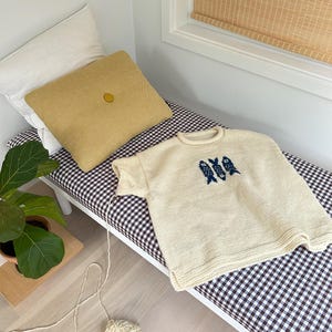 K&ouml;nnte beinhalten: Ein cremefarbener, kurz&auml;rmeliger Strickpullover mit einer dunkelblauen Grafik von drei stilisierten Fischen auf der Brust. Der Pullover hat einen gerippten Rundhalsausschnitt und Saum sowie einen Faden, der zu einem Wollkn&auml;uel f&uuml;hrt.