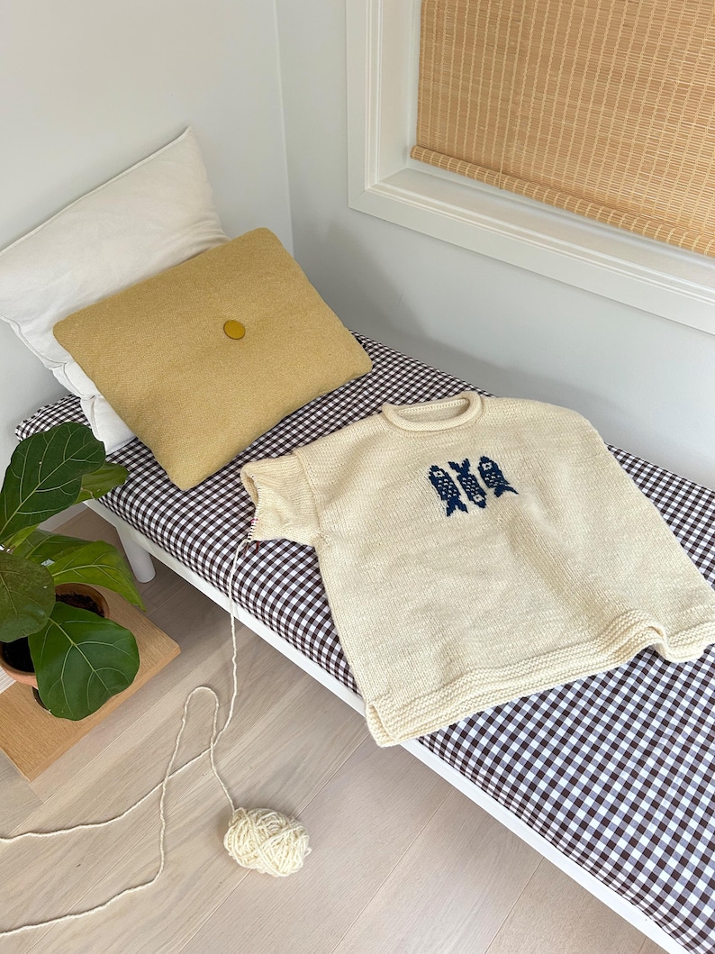 K&ouml;nnte beinhalten: Ein cremefarbener, kurz&auml;rmeliger Strickpullover mit einer dunkelblauen Grafik von drei stilisierten Fischen auf der Brust. Der Pullover hat einen gerippten Rundhalsausschnitt und Saum sowie einen Faden, der zu einem Wollkn&auml;uel f&uuml;hrt.