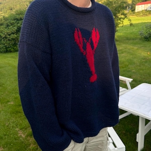 K&ouml;nnte beinhalten: Ein dunkelblauer, lang&auml;rmeliger Strickpullover mit einer auff&auml;lligen, roten Hummergrafik in der Mitte der Brust. Der Pullover hat einen gerippten Rundhalsausschnitt und eine dicke, strukturierte Strickoptik, die einen klassischen Look erzeugt.