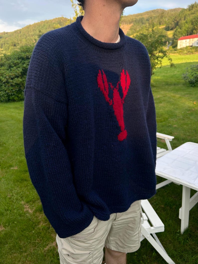 K&ouml;nnte beinhalten: Ein dunkelblauer, lang&auml;rmeliger Strickpullover mit einer auff&auml;lligen, roten Hummergrafik in der Mitte der Brust. Der Pullover hat einen gerippten Rundhalsausschnitt und eine dicke, strukturierte Strickoptik, die einen klassischen Look erzeugt.