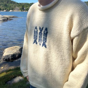 K&ouml;nnte beinhalten: Ein cremefarbener, lang&auml;rmeliger Strickpullover mit Rundhalsausschnitt und einem dunkelblauen Grafikmuster aus drei stilisierten Fischen auf der Brust. Das Kleidungsst&uuml;ck ist aus dickem, strukturiertem Garn gefertigt und hat gerippte B&uuml;ndchen.