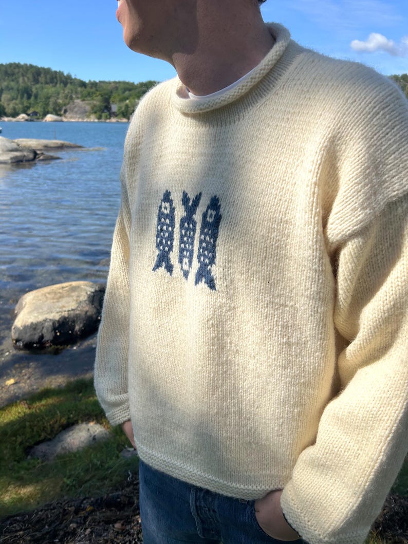 K&ouml;nnte beinhalten: Ein cremefarbener, lang&auml;rmeliger Strickpullover mit Rundhalsausschnitt und einem dunkelblauen Grafikmuster aus drei stilisierten Fischen auf der Brust. Das Kleidungsst&uuml;ck ist aus dickem, strukturiertem Garn gefertigt und hat gerippte B&uuml;ndchen.