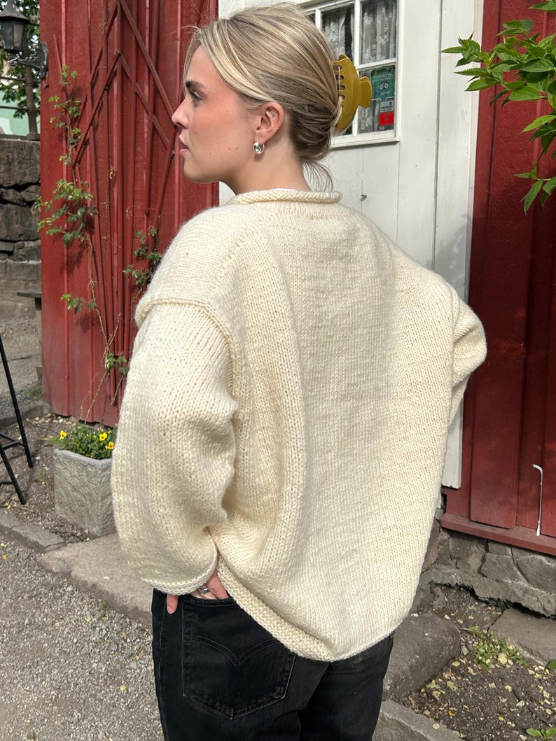 K&ouml;nnte beinhalten: Ein cremewei&szlig;er, lang&auml;rmeliger, grob gestrickter Pullover mit Rundhalsausschnitt und gerippten B&uuml;ndchen, kombiniert mit einer schwarzen Jeans. Der Pullover zeichnet sich durch eine dicke, strukturierte Garnverarbeitung aus.