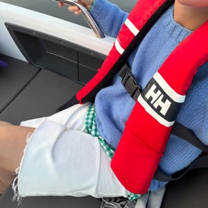 May include: A bright red life vest with white stripes and the letters HH printed in white, worn over a light blue knit jumper, a green and white checked shirt, and white denim shorts with a frayed hemline while sitting on a boat.