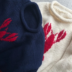 K&ouml;nnte beinhalten: Zwei Strickpullover, einer in Marineblau und einer in Creme, jeweils mit einer auff&auml;lligen roten Hummergrafik auf der Vorderseite. Beide Kleidungsst&uuml;cke haben einen Rundhalsausschnitt mit Rollkante und eine strukturierte Strickoptik.