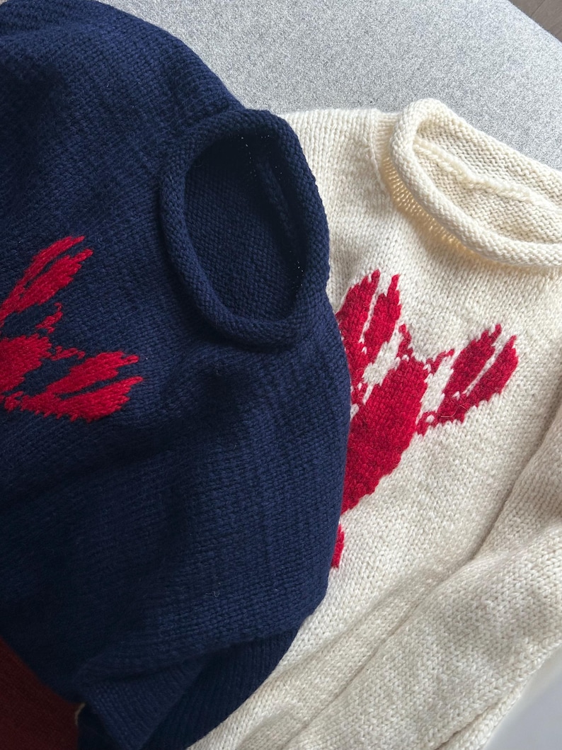 K&ouml;nnte beinhalten: Zwei Strickpullover, einer in Marineblau und einer in Creme, jeweils mit einer auff&auml;lligen roten Hummergrafik auf der Vorderseite. Beide Kleidungsst&uuml;cke haben einen Rundhalsausschnitt mit Rollkante und eine strukturierte Strickoptik.