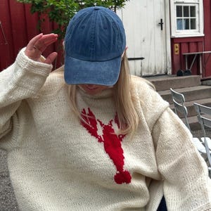 K&ouml;nnte beinhalten: Ein cremefarbener, lang&auml;rmeliger Strickpullover mit einer gro&szlig;en, kr&auml;ftig roten Hummergrafik in der Mitte der Vorderseite, kombiniert mit einer einfarbigen blauen Baseballkappe. Der Pullover hat ein dickes, strukturiertes Strickmuster.