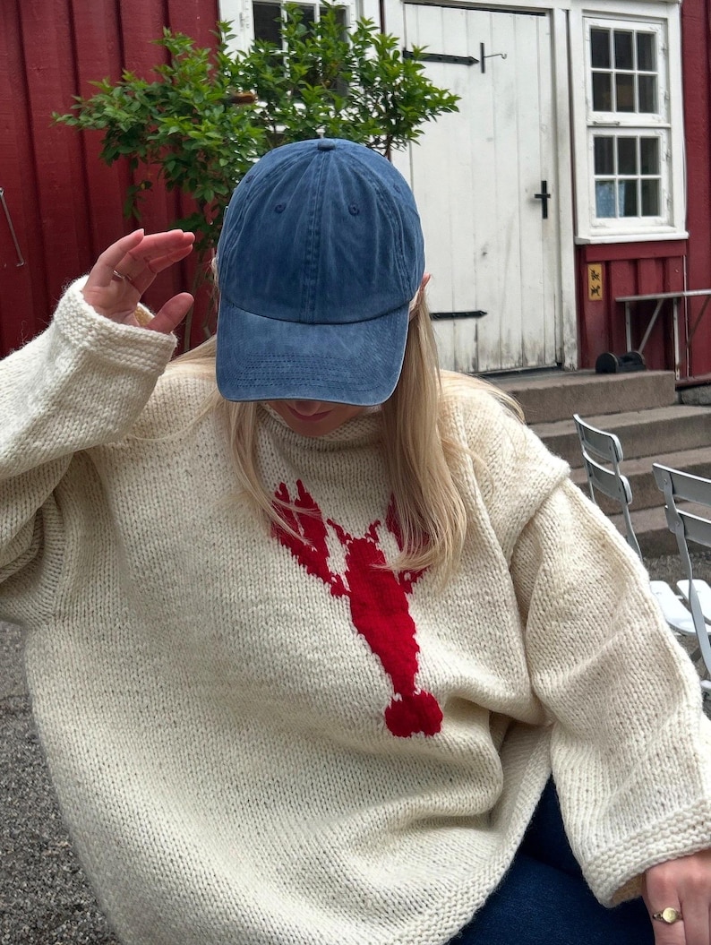 K&ouml;nnte beinhalten: Ein cremefarbener, lang&auml;rmeliger Strickpullover mit einer gro&szlig;en, kr&auml;ftig roten Hummergrafik in der Mitte der Vorderseite, kombiniert mit einer einfarbigen blauen Baseballkappe. Der Pullover hat ein dickes, strukturiertes Strickmuster.