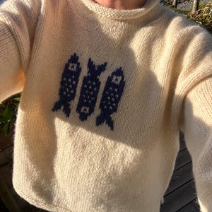 K&ouml;nnte beinhalten: Ein cremefarbener, lang&auml;rmeliger Strickpullover mit Rundhalsausschnitt und einem dunkelblauen Grafikmuster aus drei vertikalen Fischen, die mittig auf der Brust platziert sind. Der Pullover besteht aus einem dicken, strukturierten Garn.