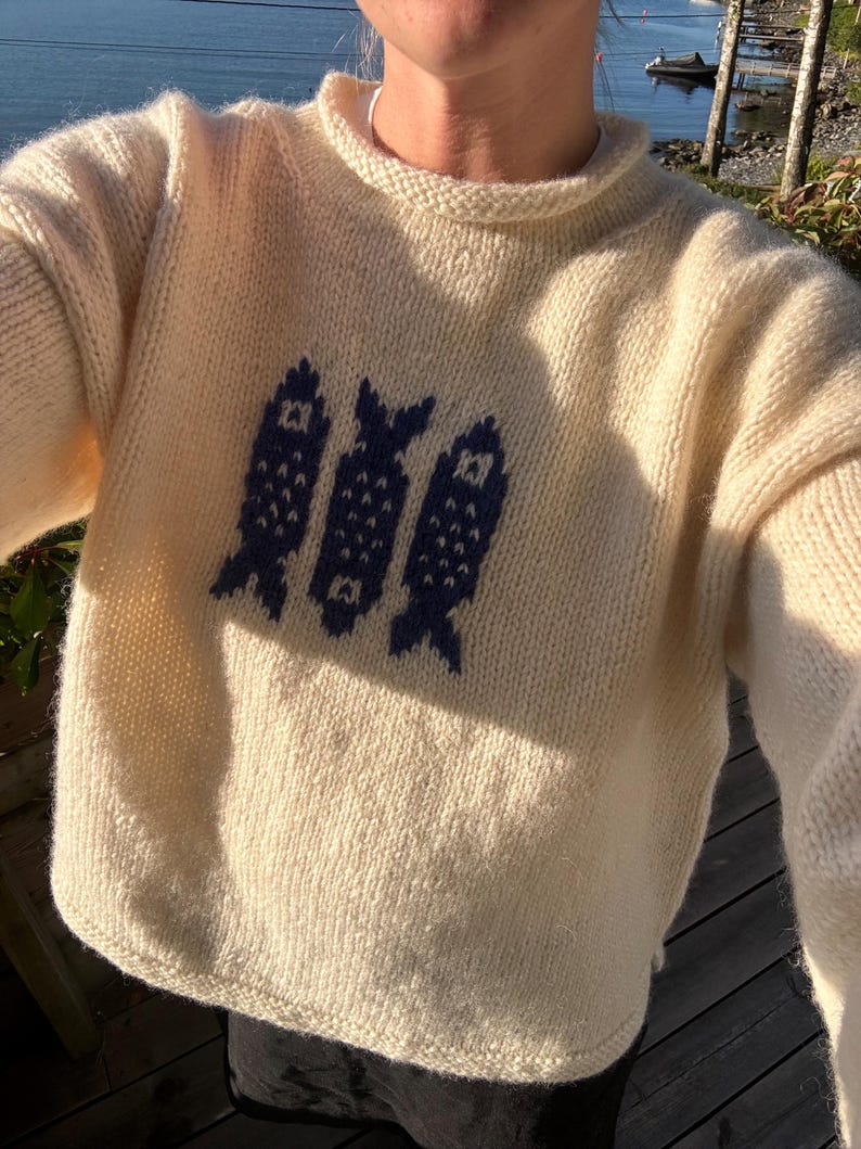 K&ouml;nnte beinhalten: Ein cremefarbener, lang&auml;rmeliger Strickpullover mit Rundhalsausschnitt und einem dunkelblauen Grafikmuster aus drei vertikalen Fischen, die mittig auf der Brust platziert sind. Der Pullover besteht aus einem dicken, strukturierten Garn.