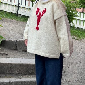 K&ouml;nnte beinhalten: Ein &uuml;bergro&szlig;er, cremefarbener Strickpullover mit einer auff&auml;lligen roten Hummergrafik auf der Vorderseite. Das Kleidungsst&uuml;ck hat einen hohen Ausschnitt, lange &Auml;rmel mit einer leichten Farbabweichung an den Au&szlig;enarmen und einen geraden Saum.
