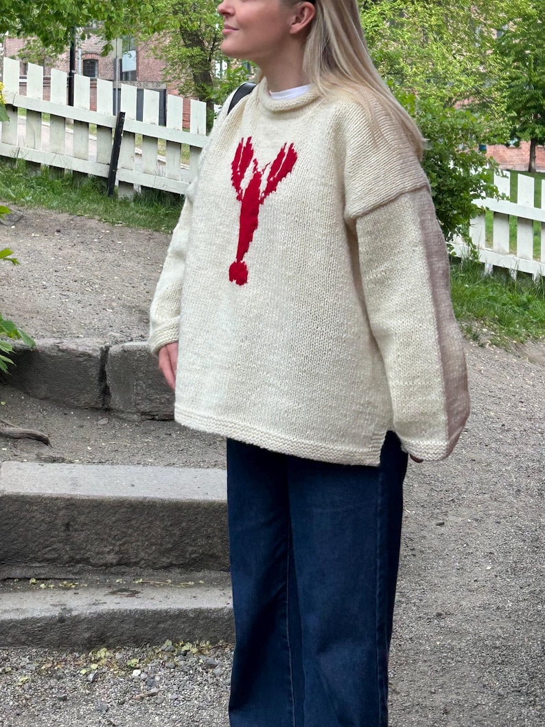 K&ouml;nnte beinhalten: Ein &uuml;bergro&szlig;er, cremefarbener Strickpullover mit einer auff&auml;lligen roten Hummergrafik auf der Vorderseite. Das Kleidungsst&uuml;ck hat einen hohen Ausschnitt, lange &Auml;rmel mit einer leichten Farbabweichung an den Au&szlig;enarmen und einen geraden Saum.